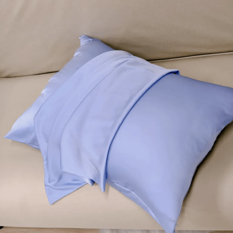 A light blue mulberry silk pillowcase draped elegantly over another matching silk pillow on a beige leather sofa.