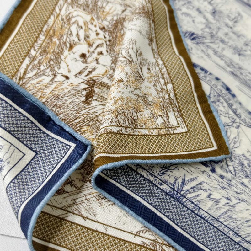 A close-up details of a folded Docsun silk-wool blend scarf, showcasing vintage nature prints in tan and navy blue with a light blue hand-rolled edge.