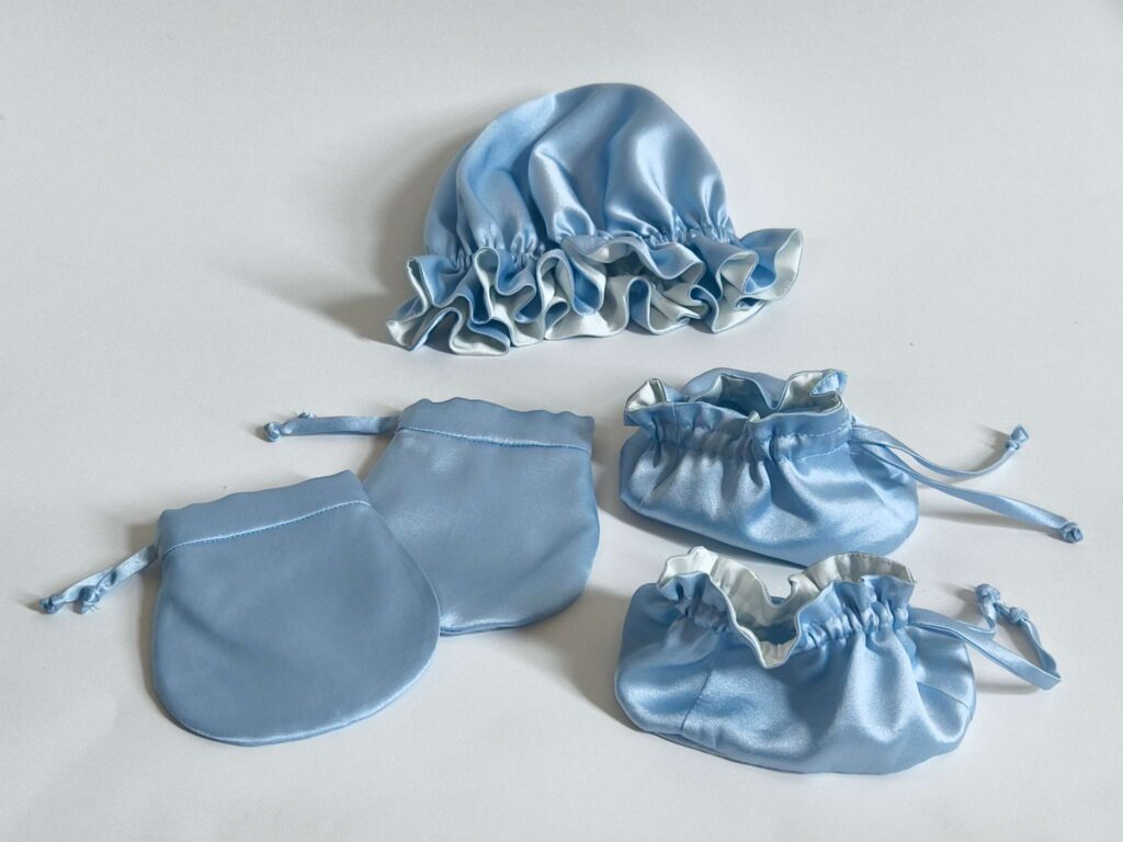 100% mulberry silk baby accessories set in soft blue, including a ruffled bonnet, mittens, and drawstring booties.