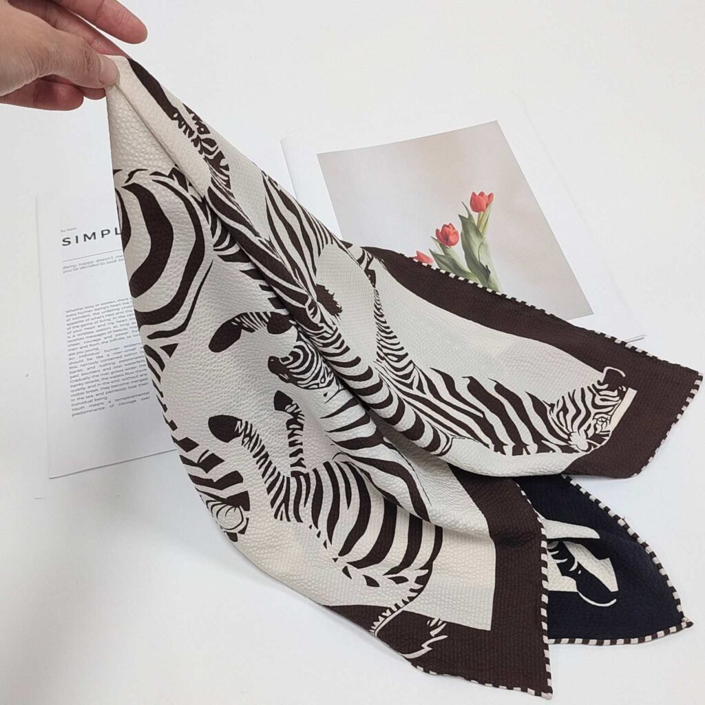 A hand holding up a Docsun 68cm mulberry silk square scarf with a black and white zebra pattern and dark border.
