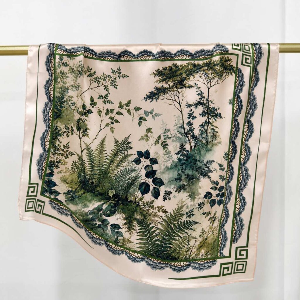 Forest botanical silk square scarf hanging on a rod, featuring an ivory beige base, green foliage print, and geometric border design.