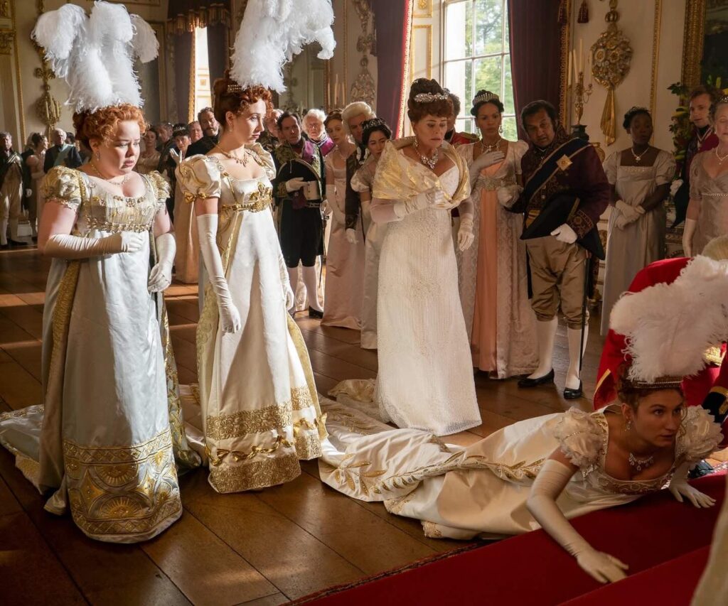 Four young women, the Featherington sisters, at a debutante ball, wearing ornate white and gold gowns and elaborate feather headdresses. Three stand together, looking worried, while the fourth lies face-down on the floor in the foreground, near a person in a detailed military-style uniform. The setting is a grand, crowded historical ballroom.