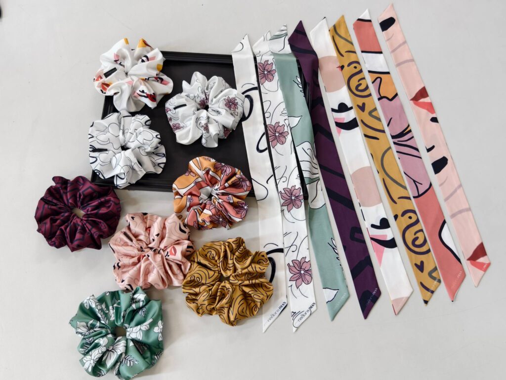 A flat-lay display of printed silk scrunchies and matching long silk ribbons, featuring floral, line-art, abstract patterns, and soft color combinations.