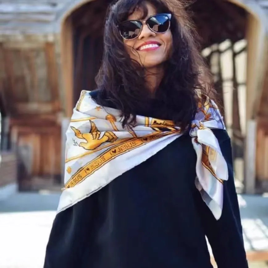 A woman wearing a white and gold printed silk scarf draped over her shoulders, styled with a black top and sunglasses for an elegant outdoor look.