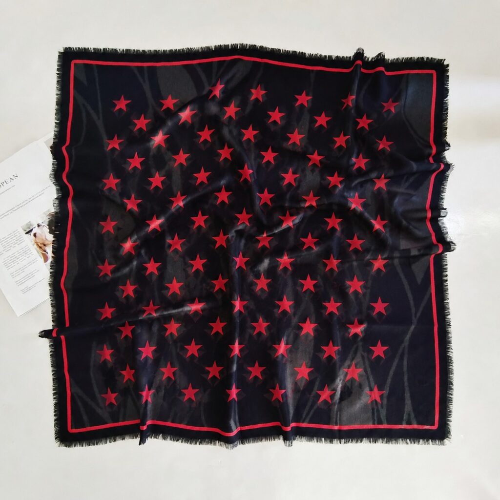 A black silk-wool blend square scarf displayed flat, featuring evenly arranged red five-pointed stars, a red border, and black fringed edges.