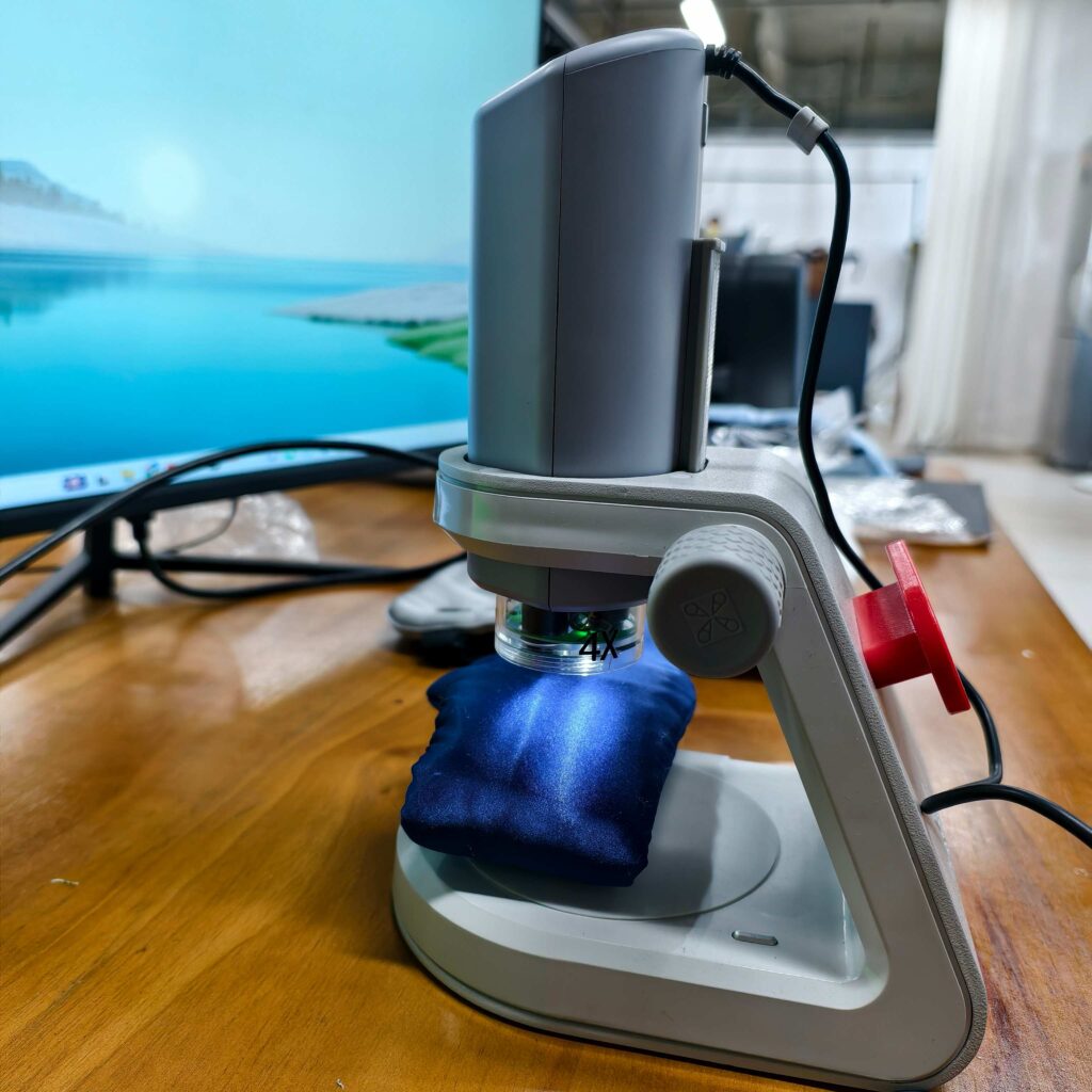 A digital microscope observing blue satin fabric at 4x magnification, showing the fabric sheen and surface condition under light.